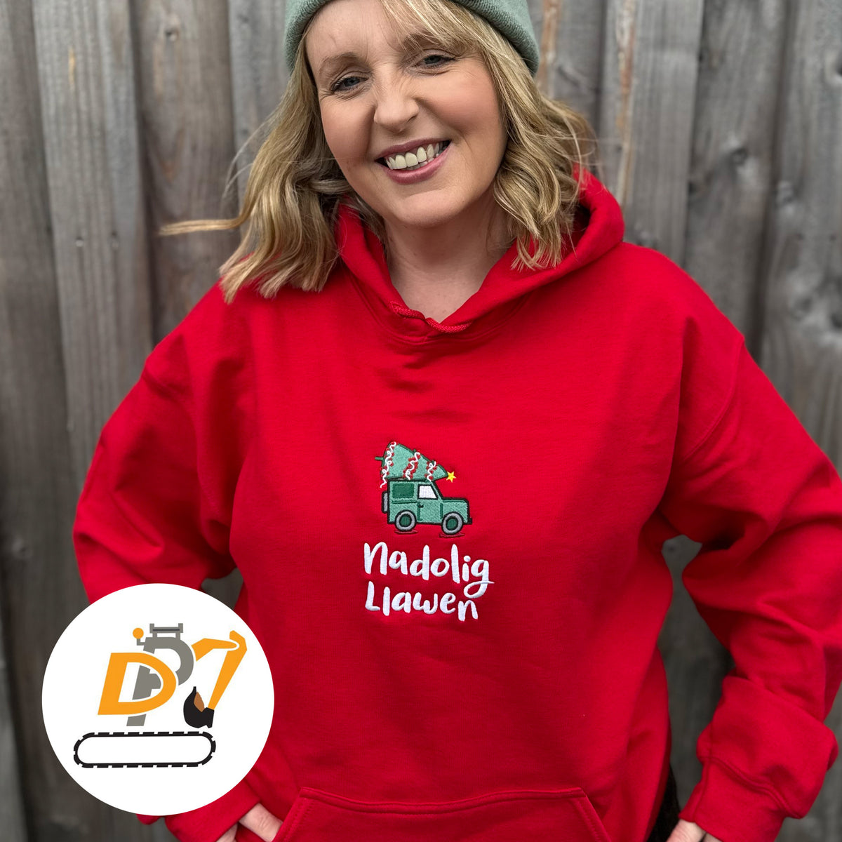 A woman wearing a bright red hoodie with a green embroidered Christmas Land Rover  graphic and the Welsh text 'Nadolig Llawen' on the front, standing against a wooden fence. A circular DPJ Foundation logo overlay is in the bottom-left corner.