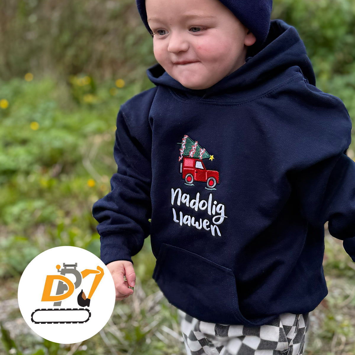 A toddler wearing a navy blue hoodie with a red Christmas tree truck graphic and the text 'Nadolig Llawer' on the front, standing outdoors in a grassy area. A circular logo watermark is visible in the bottom-left corner.