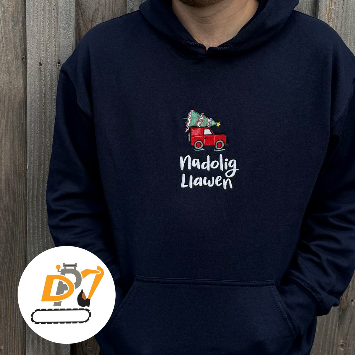 A navy blue hoodie featuring a red Land Rover carrying a Christmas tree with the Welsh text 'Nadolig Llawen' in white script