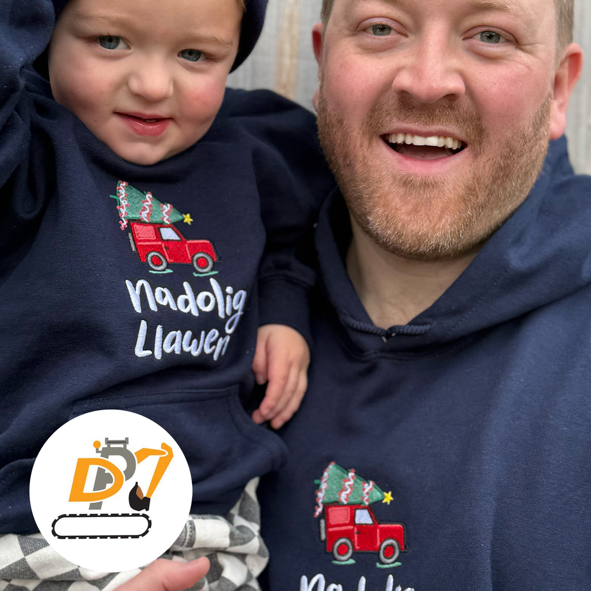 A man and a toddler wearing matching navy blue hoodies with a red Land Rover carrying a Christmas tree graphic and the Welsh text 'Nadolig Llawen' in white.