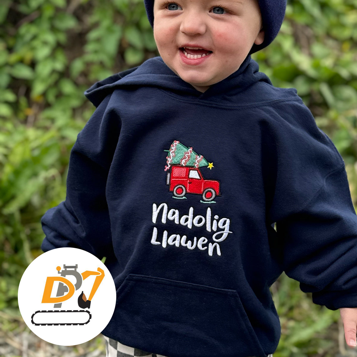 A toddler wearing a navy blue hoodie with an embroidered red Christmas tree truck graphic and the text 'Nadolig Llawen' on the front, photographed outdoors with blurred green foliage in the background. A circular logo watermark appears in the lower-left corner.