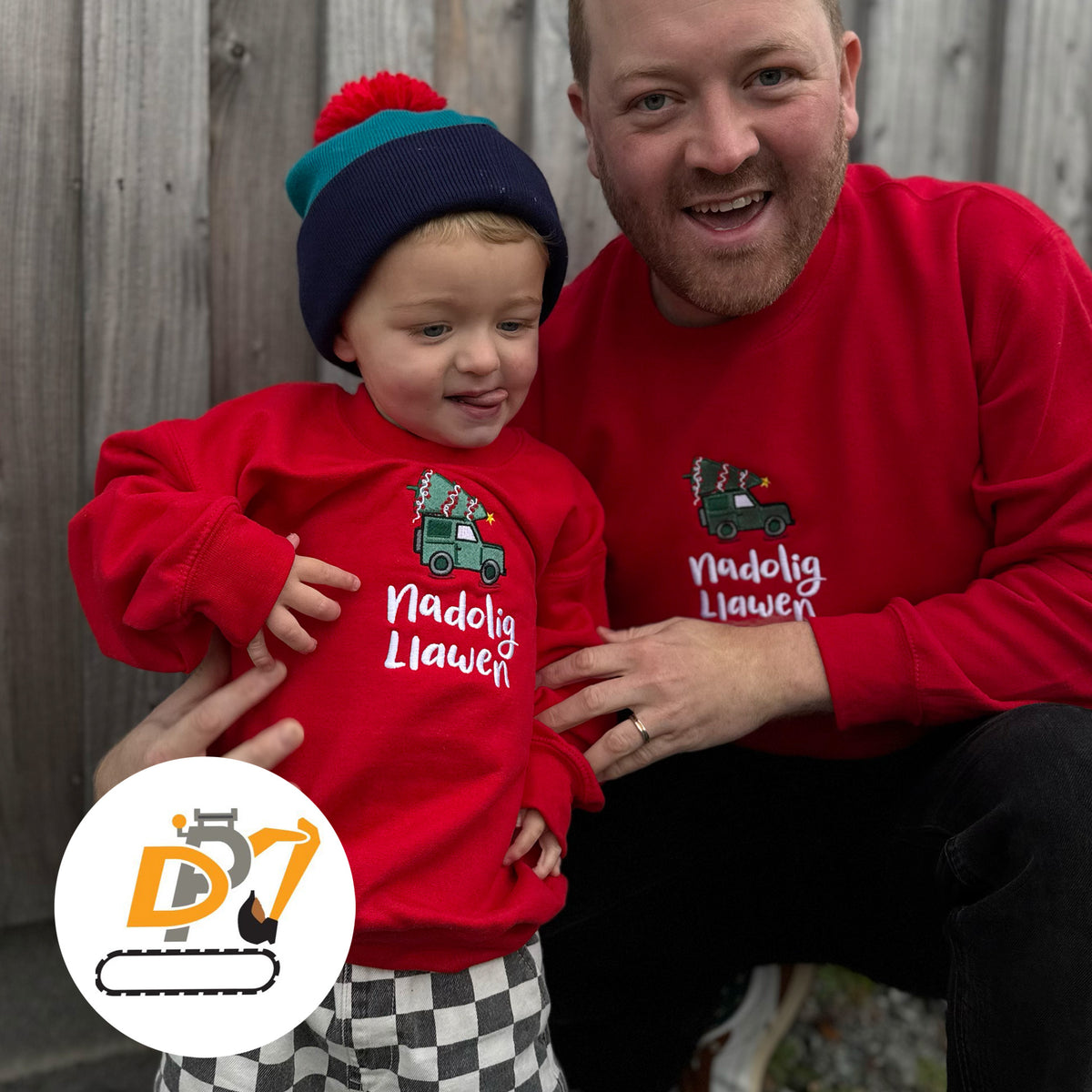 A man and a young child wearing matching red Christmas sweatshirts with a green Land Rover embroidered design and the Welsh text 'Nadolig Llawen'. 