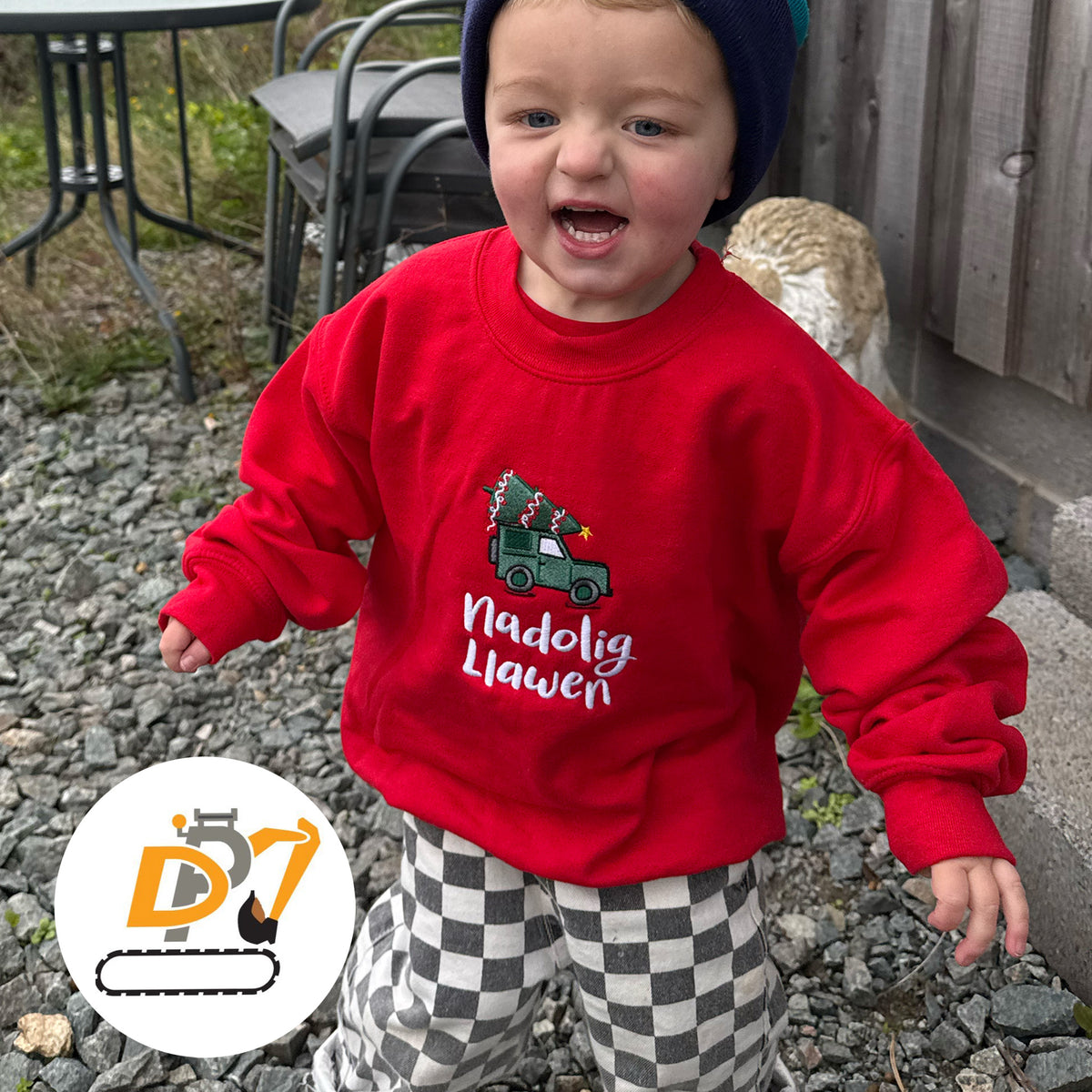 A toddler wearing a bright red Christmas jumper with a green Land Rover carrying a Christmas tree and the Welsh language text 'Nadolig Llawen' in white. 