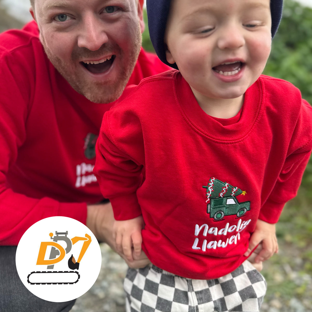 A man and a young child wearing matching red jumpers with a Christmas tree truck graphic and the Welsh text 'Nadolig Llawen' on the front