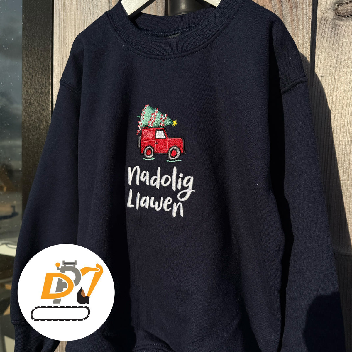 A navy blue Christmas sweatshirt hanging against a wooden background. The sweatshirt features a graphic of a red Land Rover carrying a Christmas tree above the white text 'Nadolig Llawen' (Welsh for 'Merry Christmas').